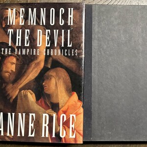 May include: A hardcover book titled "Memnoch the Devil" by Anne Rice, part of "The Vampire Chronicles" series. The cover features a Renaissance-style painting with figures in shades of brown, red, and white. The open book reveals a gray cover.