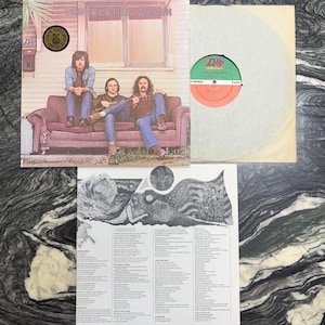 May include: An album cover featuring Crosby, Stills & Nash, with the band members seated on a purple couch. The album is accompanied by a record and a lyric sheet. The record label is green and red.
