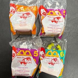 May include: Four sealed McDonald's Happy Meal toys from the film Space Jam. The toys are in clear plastic packaging with colourful cardboard featuring characters like Daffy Duck and the Tasmanian Devil. The packaging has the Warner Bros and Space Jam logos.