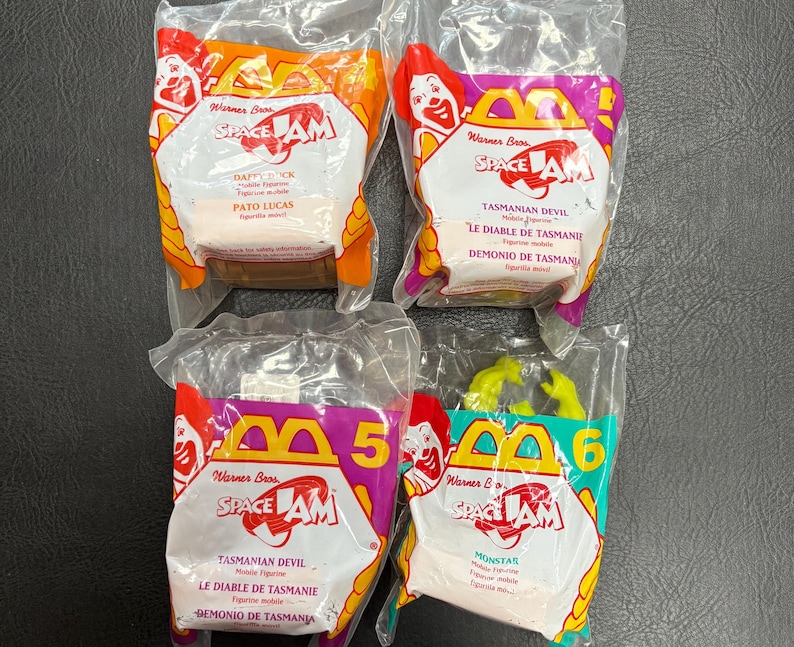 May include: Four sealed McDonald's Happy Meal toys from the film Space Jam. The toys are in clear plastic packaging with colourful cardboard featuring characters like Daffy Duck and the Tasmanian Devil. The packaging has the Warner Bros and Space Jam logos.