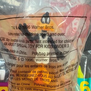 May include: A Daffy Duck toy figure in a clear plastic bag. The bag has text that reads "TM & &copy; 1996 Warner Bros." and "Safety-tested for children age 3 and over." The toy is orange, black, and white.
