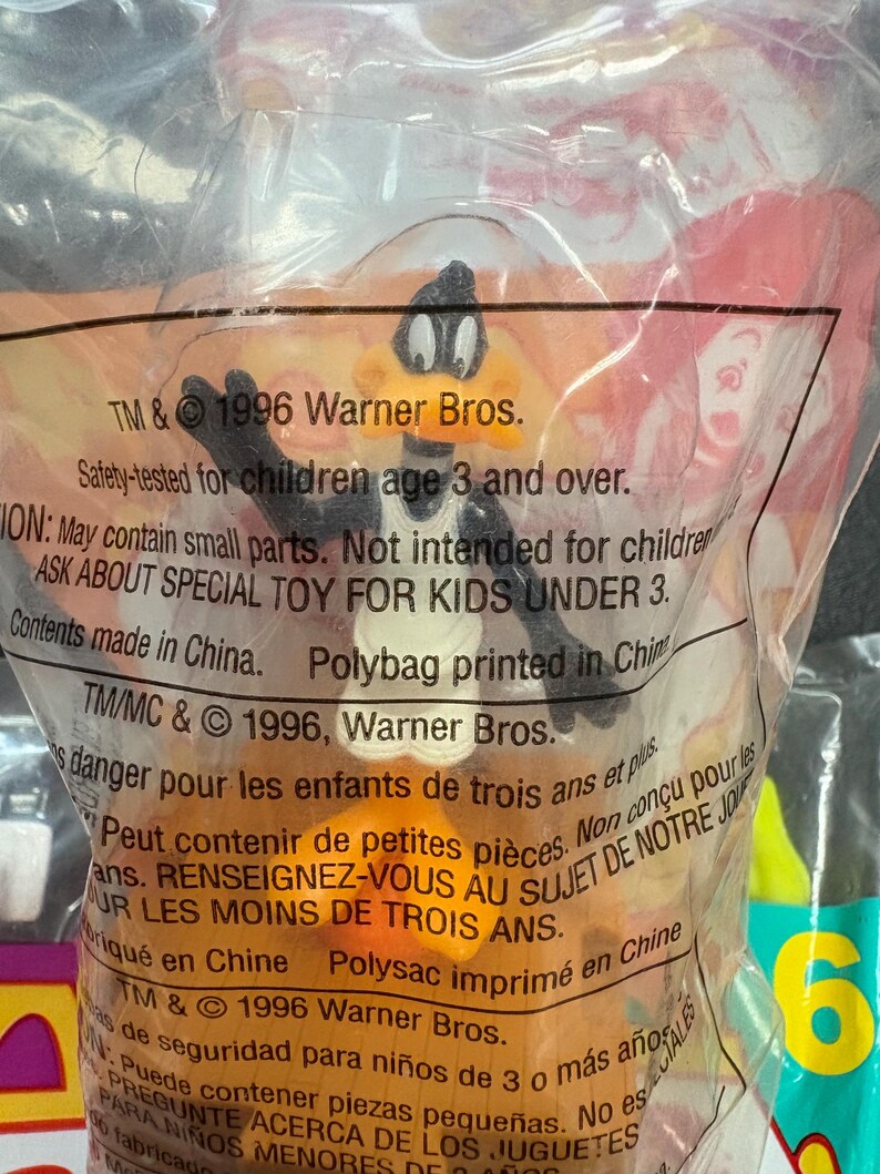 May include: A Daffy Duck toy figure in a clear plastic bag. The bag has text that reads "TM & &copy; 1996 Warner Bros." and "Safety-tested for children age 3 and over." The toy is orange, black, and white.