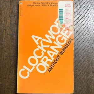 May include: A well-worn paperback copy of Anthony Burgess's "A Clockwork Orange". The book's cover is a vibrant orange, featuring the title in bold white lettering. There's a price sticker and some wear and tear visible on the top right corner.