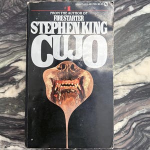 May include: A mass market paperback book titled Cujo by Stephen King. The black cover features white text and a central illustration of a dog's snout with bared teeth and saliva dripping downward. The text reads From the author of Firestarter.