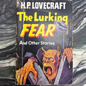 Pode incluir: Livro de bolso vintage intitulado "The Lurking Fear and Other Stories" de H.P. Lovecraft. A capa apresenta uma ilustração sombria de um monstro emergindo de um túmulo, com o título em letras amarelas e vermelhas.