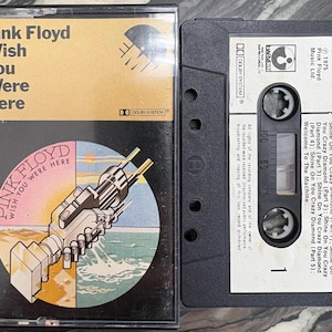 Op de afbeelding: Een vintage cassettebandje van Pink Floyd's album "Wish You Were Here". De albumhoes toont een mechanisch handdrukontwerp op een kleurrijke, gesegmenteerde achtergrond. De cassettebehuizing is zwart.