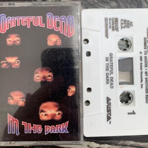 May include: A vintage cassette tape of the Grateful Dead's album "In the Dark." The album cover features the band's name in red and blue, with eyes on a black background. The white cassette tape has song titles and the Arista Records logo.