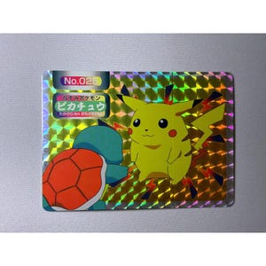 May include: A rectangular holographic trading card featuring Pikachu and Squirtle. The card displays the text No.025, Pikachu in Japanese, and stats including 0.4m and 6.0kg. The background has a gold-toned geometric pattern.