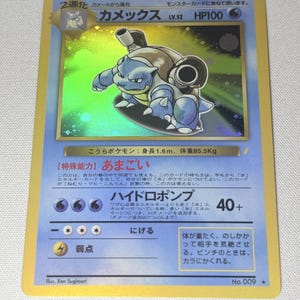 May include: A Japanese Blastoise trading card featuring a holographic illustration of the blue turtle-like creature with two cannons on its back. The card displays the name Kamex, HP100, and various Japanese text including attack details and stats.