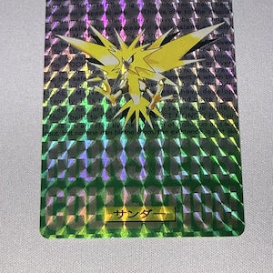 May include: A rectangular trading card featuring a holographic diamond pattern overlay. The card displays a yellow, bird-like creature with sharp wings and a spiky tail. Text includes POCKET MONSTER, J145, MONSTER COLLECTION, and Japanese characters.