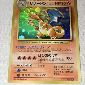 May include: A Japanese Charizard trading card featuring a holographic illustration of the orange dragon-like creature with blue wings, set against a green and black background. The card displays HP 120, fire energy symbols, and Japanese text.