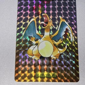 May include: A rectangular trading card featuring a holographic, checkered pattern background with the orange, dragon-like creature Charizard in the center. The card displays the text POCKET MONSTER, MONSTERS COLLECTION, and the Japanese characters リザードン.