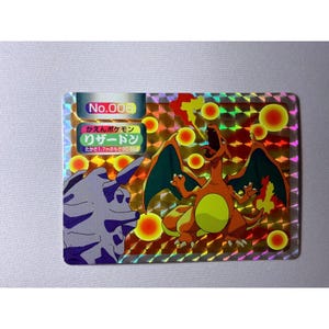 May include: A rectangular holographic trading card featuring a roaring orange dragon-like creature with teal wings and a yellow belly, alongside a grey rock-like figure. The card displays the text No.006, kaen pokemon, rizadon, and 1.7m 90.5kg.