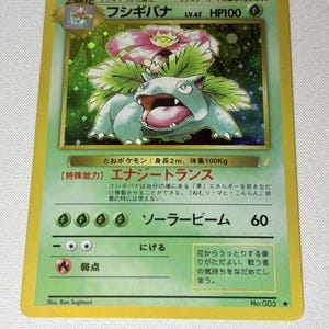 May include: A Japanese Venusaur trading card featuring a green creature with a large flower on its back. The card displays the name Fushigibana, HP100, and text including Energy Trans and Solar Beam. It has a yellow border and a holographic illustration.