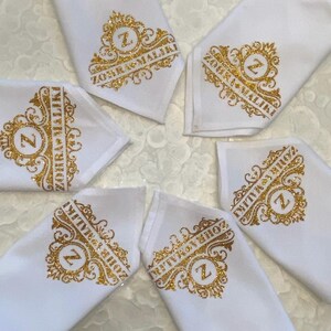 Op de afbeelding: Zes witte stoffen servetten met gouden glitterletters. Elk servet heeft een ander ontwerp met een gouden glitterletter "Z" en de tekst "ZOHRA GMAIL.LI" of "MITRVA.BIZ".