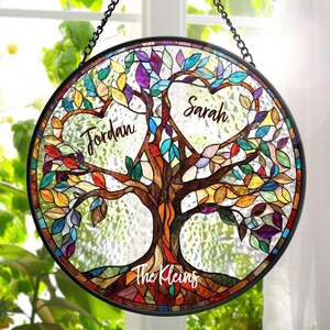 May include: Round stained glass window art featuring a tree design with colorful leaves. The names "Jordan" and "Sarah" are written within heart-shaped spaces, with "The Kleins" at the base of the tree. The artwork is framed in black and hangs from a chain.