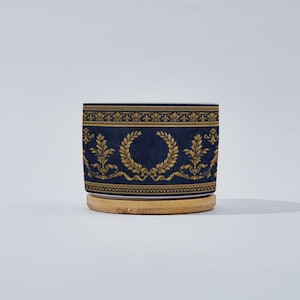 May include: A cylindrical ceramic container with a navy blue background and gold decorative elements. The design includes a laurel wreath and ornate floral patterns. The container sits on a round, light brown wooden base.