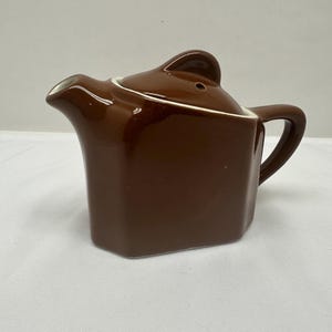 May include: A brown ceramic teapot with a square body and a curved handle. The lid has a small, rounded handle. The spout is white on the inside. The teapot is designed with a glossy finish.