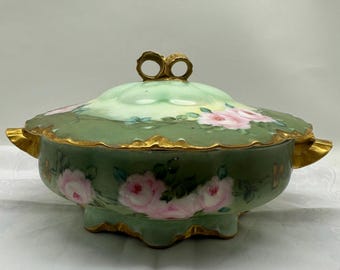 May include: A vintage, lidded serving dish with a green base and lid, adorned with pink rose floral designs. The dish features gold trim and handles, and a decorative gold bow-shaped handle on the lid. A classic piece for serving.