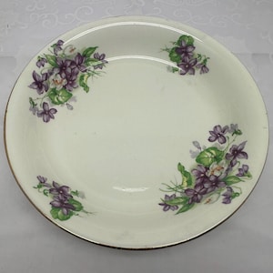 May include: Cream-colored, round ceramic plate with a gold rim. The plate is decorated with clusters of purple violets and green leaves. The floral design is arranged around the edges of the plate.