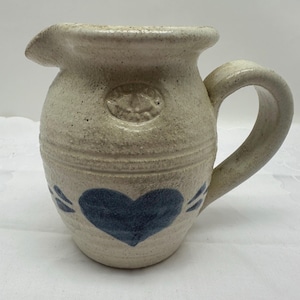 May include: A stoneware pitcher with a cream-colored speckled finish. It features a blue heart design and a handle. The pitcher has a spout for pouring and an embossed design near the top. A charming addition to any kitchen.