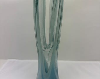 May include: A tall, elegant glass vase with a unique, organic shape. The vase features a blend of blue and clear glass, creating a gradient effect. The base is a darker shade of blue, transitioning to a lighter, almost transparent top.