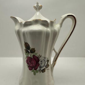 May include: White porcelain coffee pot with a lid, featuring a floral design of red and white roses with green leaves. The handle and rim are accented with gold trim. The pot has a fluted body and a decorative finial on the lid.