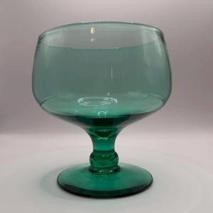 May include: A large, translucent teal-colored glass goblet featuring a wide, rounded bowl, a short, thick stem with a central decorative knob, and a circular base. The glass has a smooth, polished finish and a consistent deep green-blue hue.