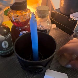 Könnte beinhalten: Eine schlanke, hellblaue Stabkerze steht aufrecht in einem schwarzen, runden Kerzenhalter. Die Kerze ist mit kleinen getrockneten Kräutern bestreut. In der Nähe befinden sich eine bärenförmige Honigflasche, ein Glas mit Korken und Pfefferminzöl.