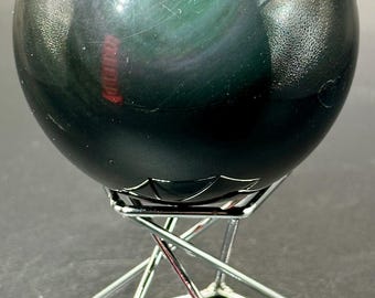May include: A dark, iridescent obsidian sphere, resting on a silver metal stand. The sphere has a polished surface with hints of green and purple. The stand is a geometric design with a crisscross pattern.