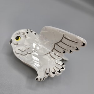 May include: A white owl-shaped hair clip with a pearlescent finish. The clip features black wing details and yellow eyes. The owl is depicted with its wings spread, giving it a dynamic appearance. The clip is designed to hold hair in place.