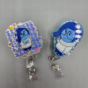 May include: Two blue and silver glitter badge holders featuring the character Sadness from the movie Inside Out. The character is wearing a white sweater and blue pants. The badge holders have clear plastic retractable reels.