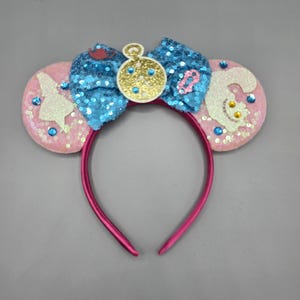 May include: Pink sequined mouse ears headband with a blue sequined bow and gold clock charm. The ears feature Alice in Wonderland characters. The headband is a bright pink color.