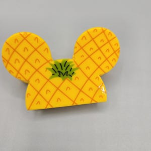 May include: A yellow pineapple-shaped hair clip with green leaves on top. The clip is designed in the shape of Mickey Mouse ears.