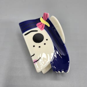 May include: A ceramic brooch depicting a stylized face. The face is white with a black eye, pink lips, and three black freckles. The hair is dark blue with a pink and yellow bow.