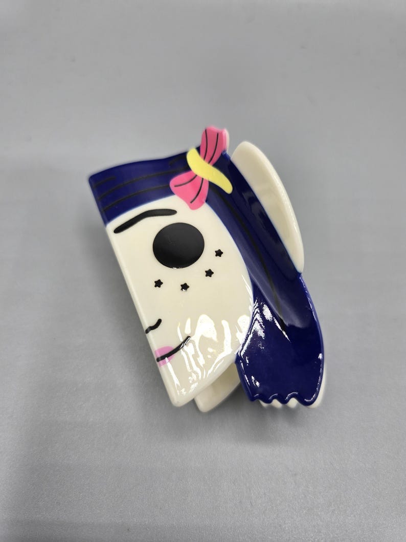 May include: A ceramic brooch depicting a stylized face. The face is white with a black eye, pink lips, and three black freckles. The hair is dark blue with a pink and yellow bow.