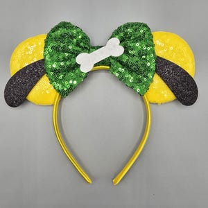 May include: A yellow headband with dog ears and a green sequined bow. The ears are yellow with black tips, and the bow features a white bone-shaped accent. This playful accessory is perfect for themed parties or costumes.