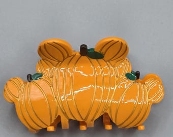 May include: Orange pumpkin-shaped hair clip with three pumpkins. Each pumpkin has a green leaf and a brown stem. The clip is made of shiny plastic and has gold-colored lines. The clip is designed to hold hair in place.