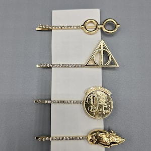 May include: Four gold-toned hair clips with rhinestone accents. The clips feature iconic shapes: round glasses, a triangle with a circle, a coin with the number 9 3/4, and a train. The clips are displayed on a white card.