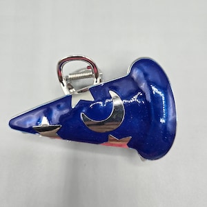 May include: A blue and silver hair clip in the shape of a wizard's hat. The clip features silver stars and a crescent moon design. The hat has a shiny, glossy finish and a silver metal clip.