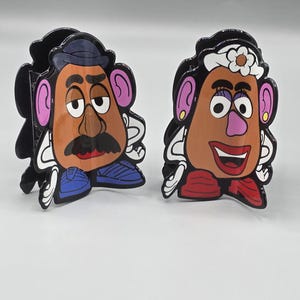 May include: Two cartoon-style character cutouts, one male and one female. The male has a brown face, black mustache, and blue shoes. The female has a white flower in her hair and a red bow. Both have black outlines.