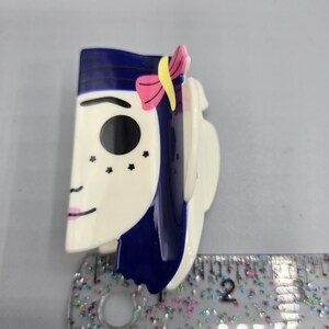 May include: A decorative hair clip featuring a stylized face with blue hair, a pink bow, and black star-shaped freckles. The clip is primarily white with blue and pink accents. The clip is approximately 5 cm long.