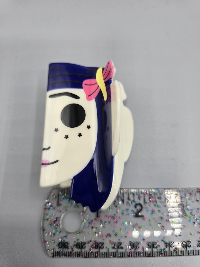May include: A decorative hair clip featuring a stylized face with blue hair, a pink bow, and black star-shaped freckles. The clip is primarily white with blue and pink accents. The clip is approximately 5 cm long.