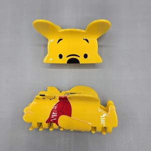 May include: Two yellow hair clips featuring a cartoon bear design. One clip is a face, the other is a side view of the bear with a red shirt. The clips are made of plastic and have a glossy finish.