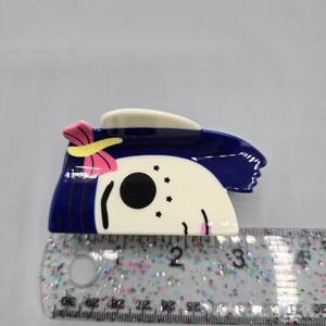 May include: A hair clip with a cartoon character design. The clip has a white face with black eyes and a blue hair design. A pink bow with a yellow center is on the side. The clip measures approximately 7.6 cm in length.