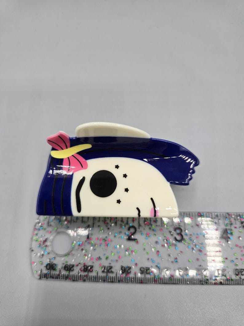 May include: A hair clip with a cartoon character design. The clip has a white face with black eyes and a blue hair design. A pink bow with a yellow center is on the side. The clip measures approximately 7.6 cm in length.