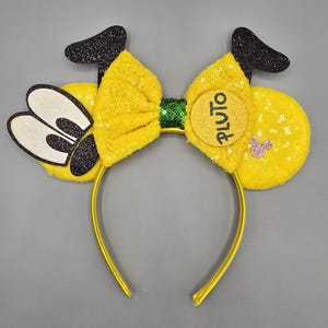 May include: Yellow headband with a large yellow bow and black glitter ears. The ears have a white face with black eyes. A yellow circle with the word "PLUTO" in black letters is attached to the bow.