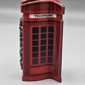 May include: A red telephone booth-shaped decorative item with the word "TELEPHONE" in white. The booth has a black grid pattern and a black chain on the side. The item is a souvenir or collectible.