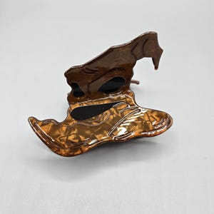 May include: A brown and gold witch hat hair clip. The clip has a shiny, textured finish with a dark brown top and a lighter, patterned brim. The hat is a decorative accessory.