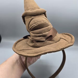 May include: A brown Sorting Hat headband with a wide brim and a tall, pointed crown. The hat features a brown ribbon and is made of a soft, textured fabric. The headband is brown.